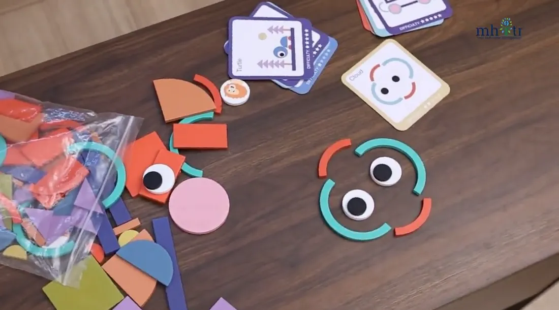 Colourful shape pieces and picture cards from the kit laid out on a table, with two pieces arranged into a face